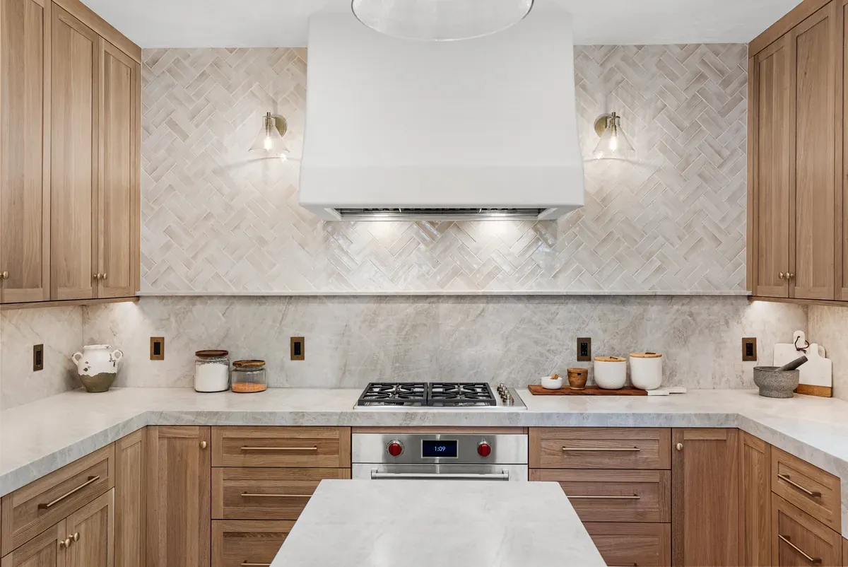 Remodeled kitchen range wall with white oak cabinets, marble herringbone backsplash and custom hood