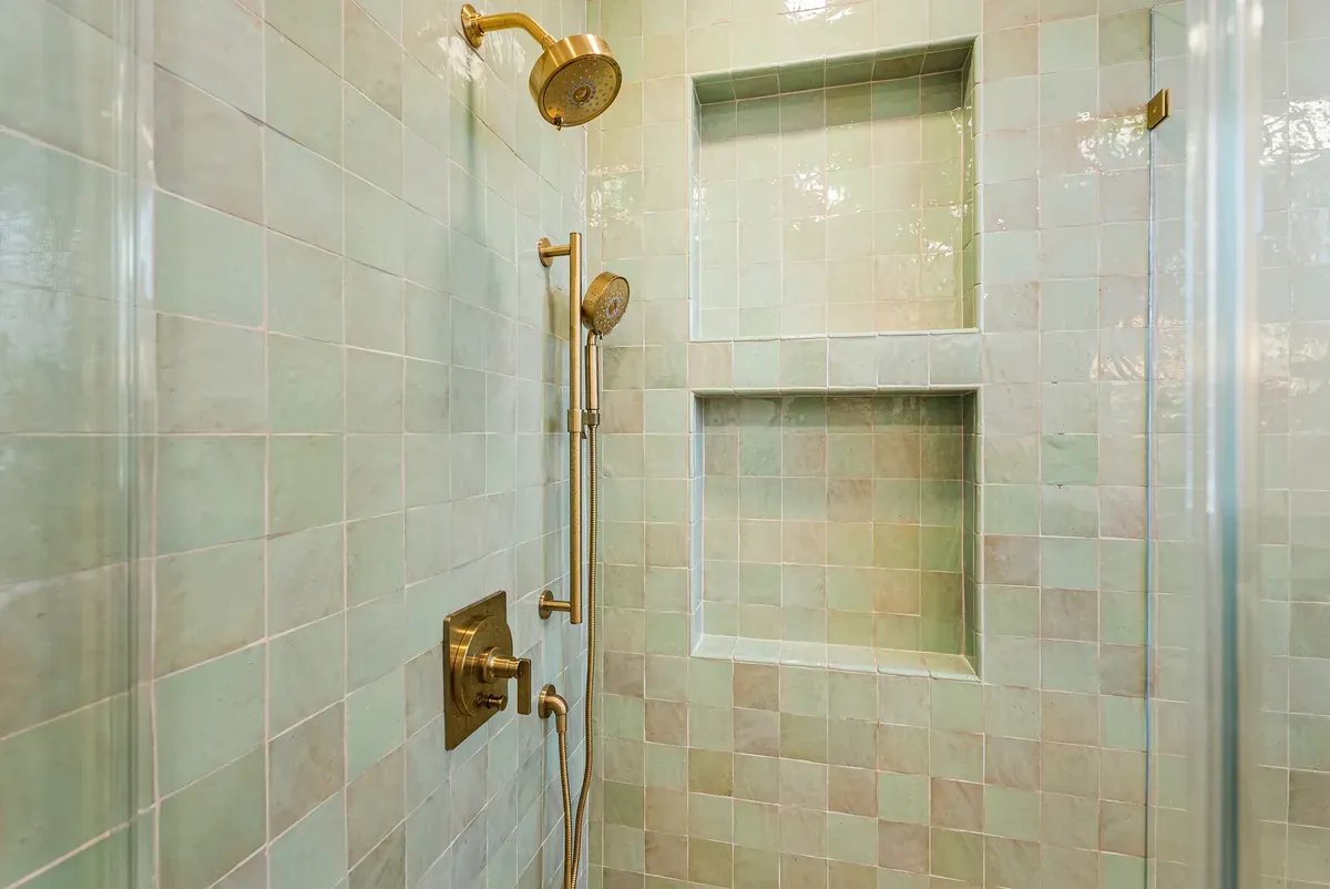 Shower interior in green zellige tile with two recessed niches, brass rain head and handheld, behind glass