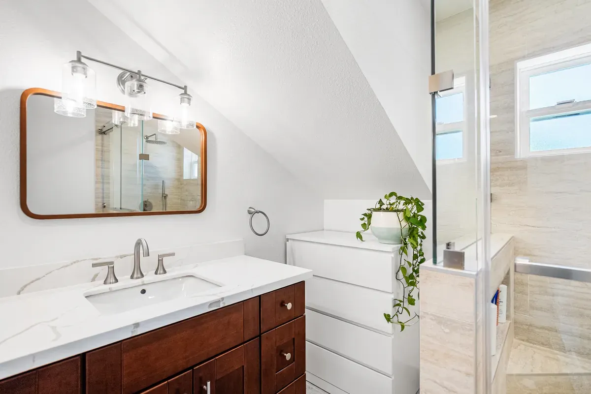 Bath vanity with marble top, dark wood cabinet and framed mirror under a sloped ceiling